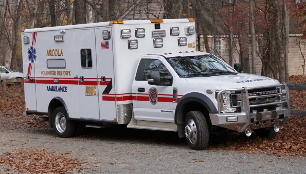 A white Ancola Volunteer Fire Dept. ambulance with red and gold stripes is parked on a gravel lot among fallen leaves and trees, with a building in the background.