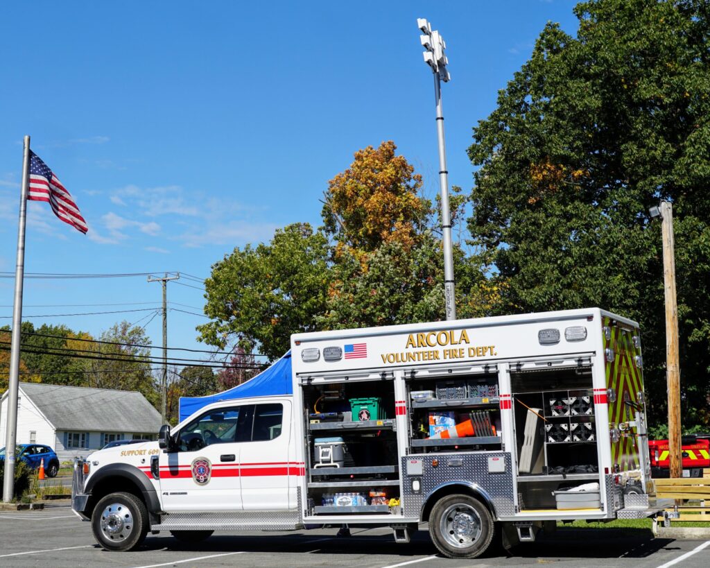 A white Arcola Volunteer Fire Dept. emergency vehicle with compartments open is parked outdoors near trees, a building, and a flagpole with the American flag. Equipment and tools are visible inside the truck.