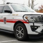 A white SUV with red stripes and emergency lights, marked with a fire department emblem and labeled “SERV 609,” is parked in a lot near a business sign and trees.