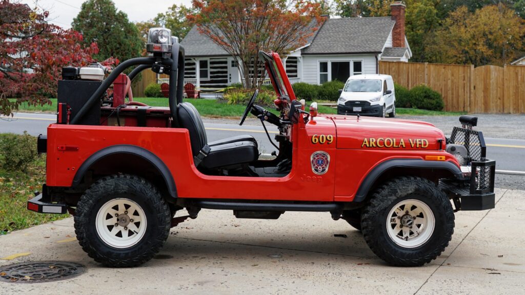 A red Arcola Fire Department Jeep with off-road tires is parked on a driveway. The vehicle has emergency lights and fire department markings. A house and a white van are visible in the background.