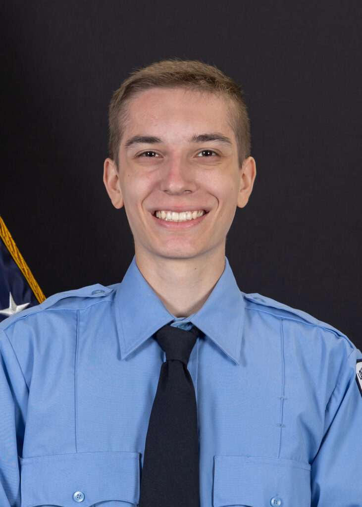 A person in a blue uniform shirt and black tie smiles at the camera, standing in front of a dark background with part of an American flag visible on the left.