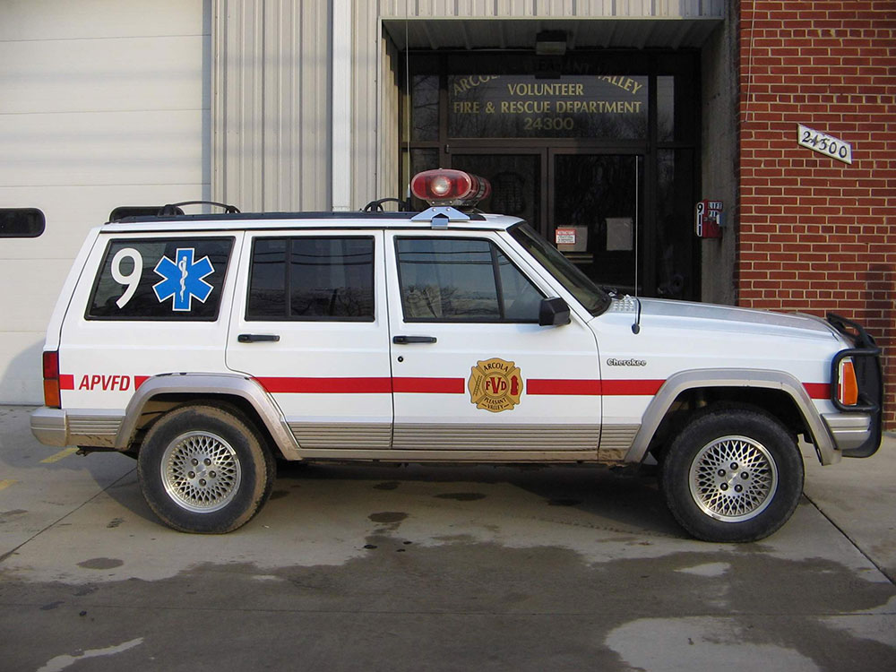 A white emergency vehicle with a red stripe, medical symbol, and fire department badge is parked outside a fire and rescue building.