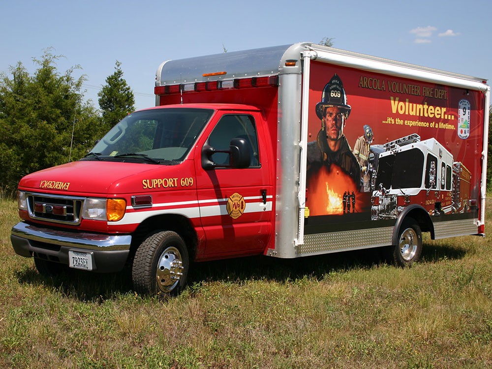 A red fire department support truck with a mural showing a firefighter, fire truck, and flames, parked on grass under a blue sky. The words “Volunteer… it’s the experience of a lifetime” are written on the side.