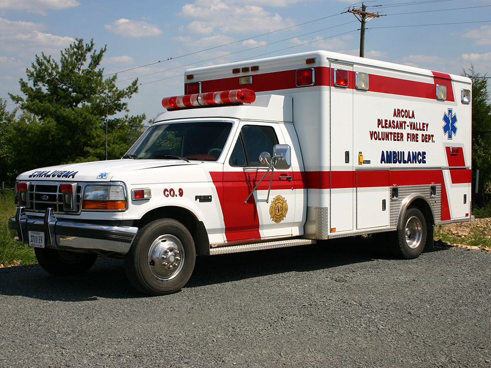 A white and red ambulance with emergency lights is parked on gravel. It has “AMBULANCE” and fire department details on the side. Green trees and a utility pole are visible in the background under a blue sky.