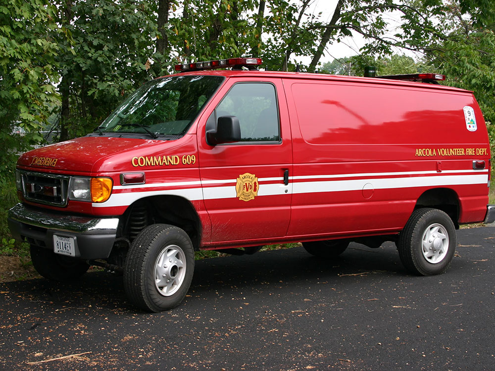 A red Arcola Volunteer Fire Department command van with white stripes is parked on a paved surface near green trees.