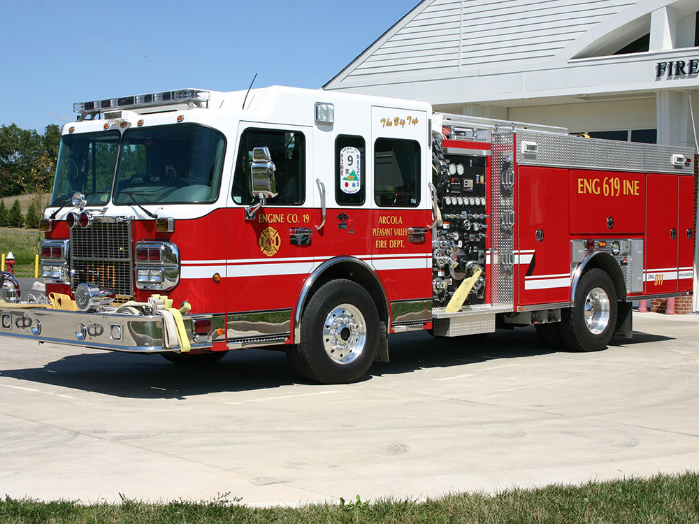 A bright red fire truck with white accents is parked outside a fire station on a sunny day. The truck has several hoses and equipment compartments visible on the side.