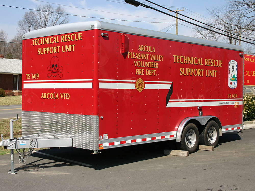 A bright red trailer labeled “Technical Rescue Support Unit” and “Arcola Pleasant Valley Volunteer Fire Dept.” is parked on pavement near a road, with trees and a brick building in the background.