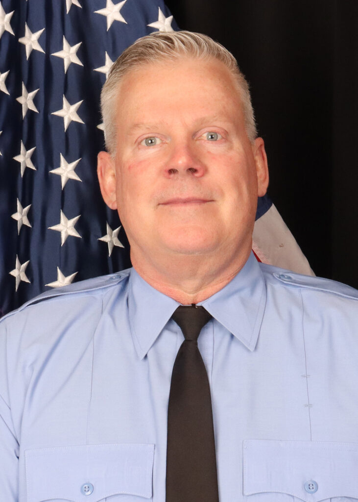 A man in a light blue uniform shirt and black tie stands in front of an American flag, smiling slightly at the camera.