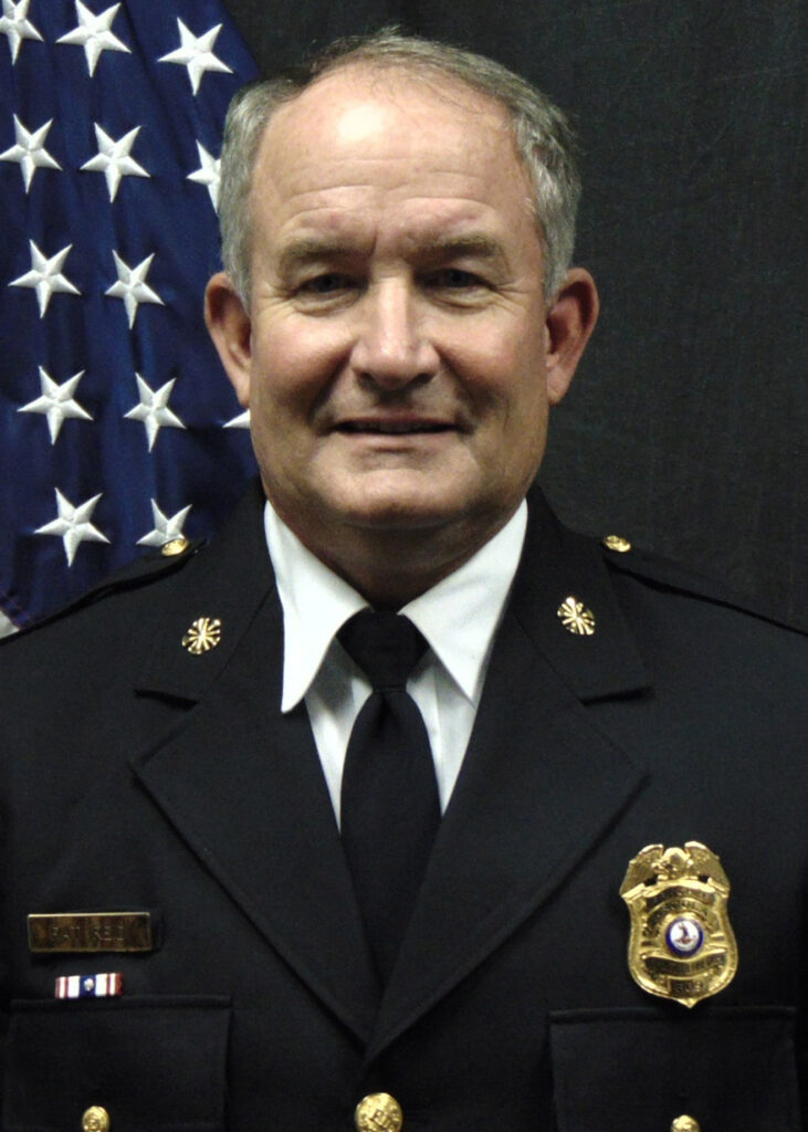 A man in a formal police uniform stands in front of a U.S. flag, facing the camera and smiling slightly. He wears a dark suit jacket with a badge, pins, and a black tie.