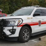 A white emergency response SUV with red stripes, official seal, and emergency lights is parked next to a curb. The front bumper has a grille guard, and trees and buildings are visible in the background.