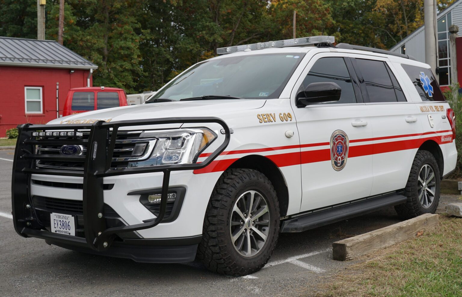 A white emergency response SUV with red stripes and a fire department emblem is parked next to a wooden barrier in a lot, with trees and a red building in the background.