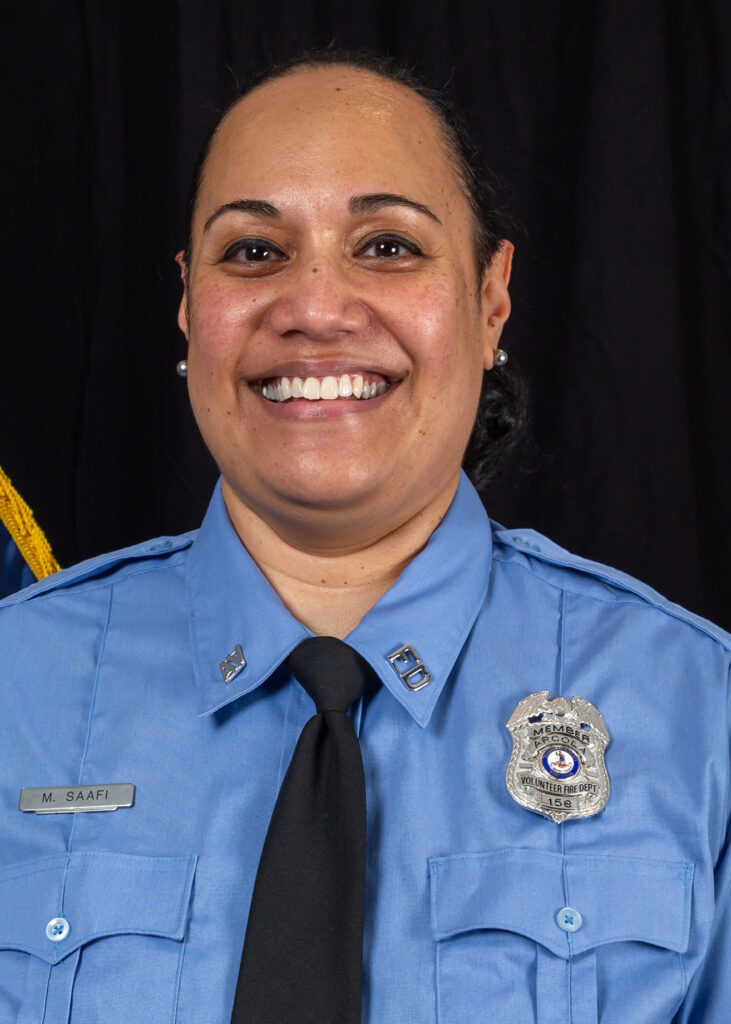 A smiling woman in a blue uniform with a tie and badge, standing in front of a black background. She has dark hair pulled back and is wearing pearl earrings.