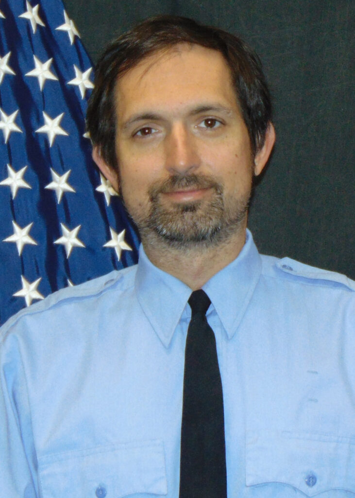 A man with short dark hair and a beard wearing a light blue uniform shirt and black tie stands in front of a U.S. flag with white stars.
