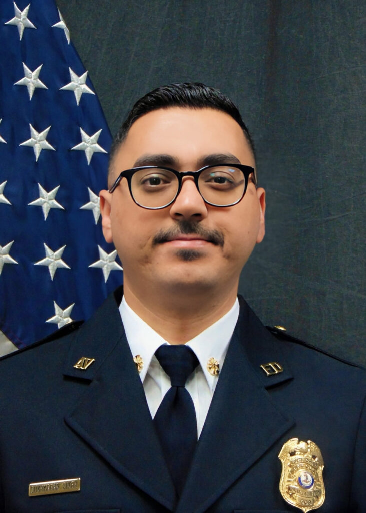 A person in a police uniform with glasses stands in front of an American flag. Their uniform displays a badge, nametag, and stars on the collar, indicating a high rank.