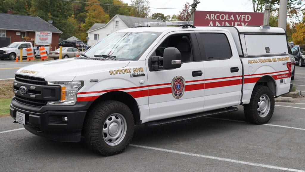 A white pickup truck with red stripes, marked “Arcola Volunteer Fire Department” and “Support 609B,” is parked in front of a building labeled “ARCOLA VFD ANNEX.”.