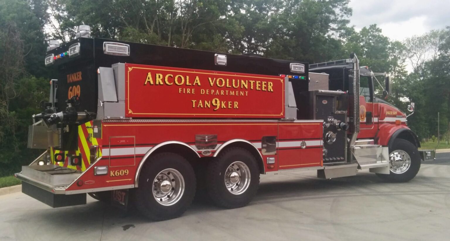 A red Arcola Volunteer Fire Department tanker truck, labeled “Tanker 609,” is parked on a paved surface with trees in the background.