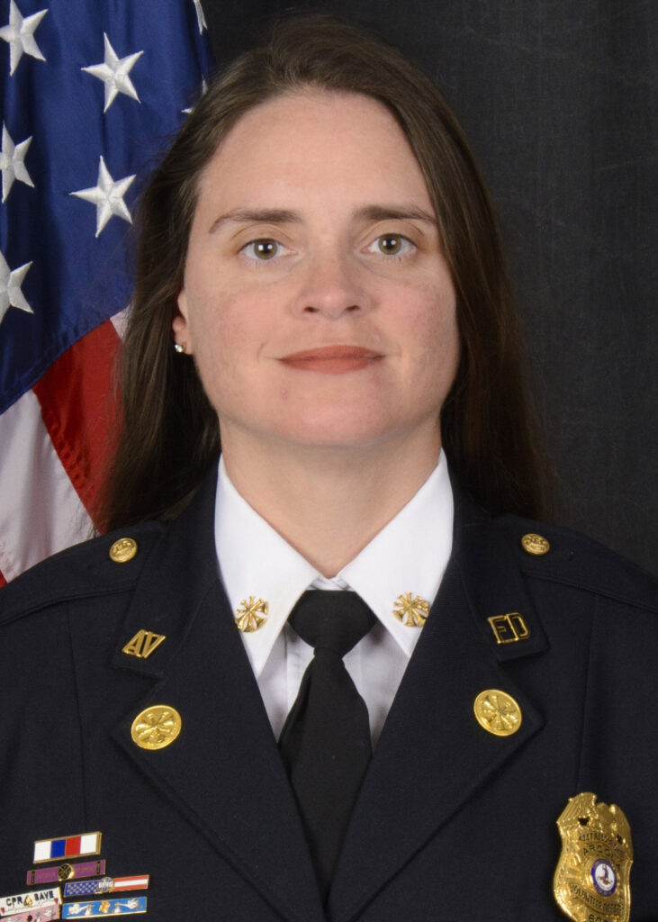 A woman in a formal dark uniform with badges and pins stands in front of an American flag, looking at the camera with a neutral expression.