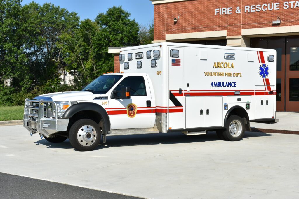 A white Arcola Volunteer Fire Dept. ambulance is parked outside a fire and rescue station on a sunny day.