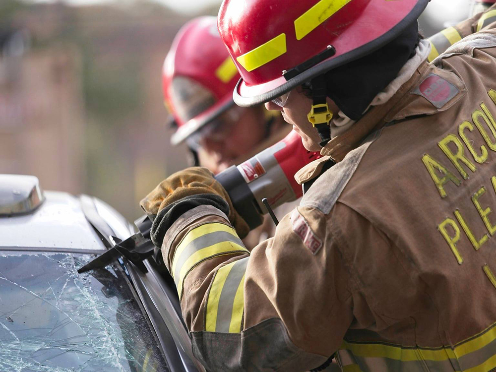 Two firefighters in helmets and protective gear use a tool to cut through the windshield of a damaged vehicle during a rescue operation.
