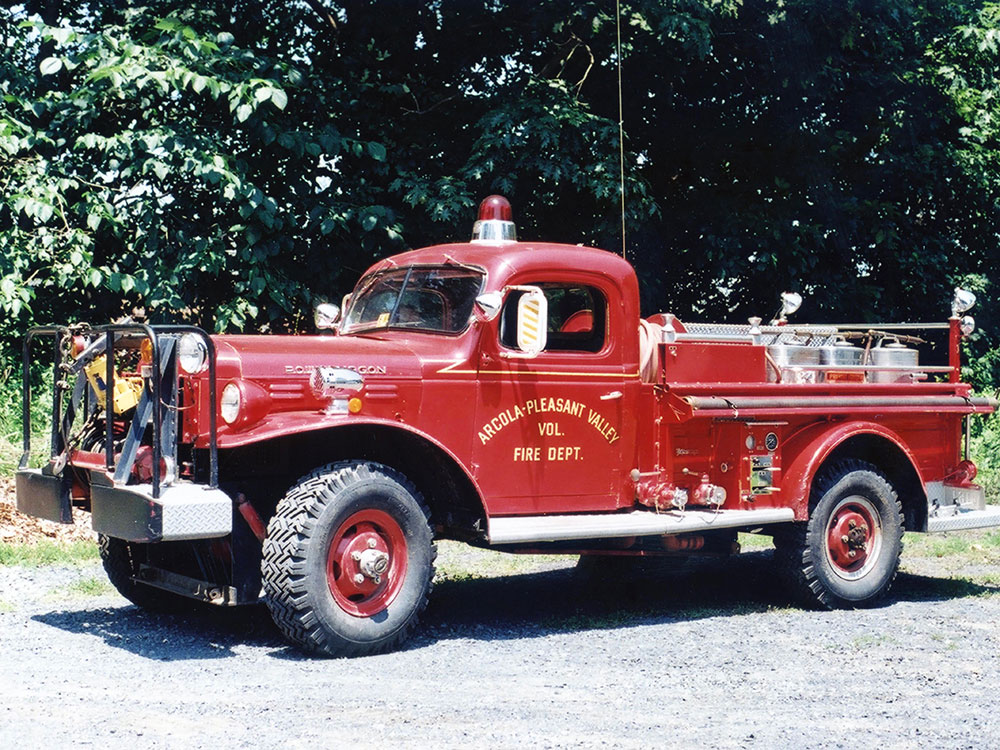 A vintage red fire truck from the Arcola-Pleasant Valley Volunteer Fire Department is parked on a gravel surface, with green trees in the background.