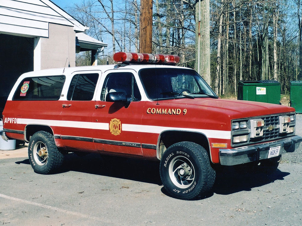 A red and white fire command SUV marked “COMMAND 9” is parked outdoors near a building and green dumpsters, surrounded by leafless trees.
