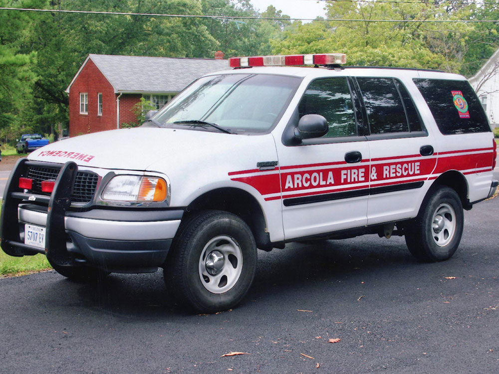 A white SUV with red “Arcola Fire & Rescue” lettering and emergency lights is parked on a driveway. A brick house and trees are in the background.