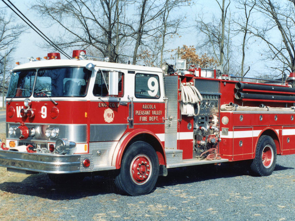 A red and white fire truck labeled “9” and “Arcola Pleasant Valley Fire Dept.” is parked on a gravel road with leafless trees in the background.