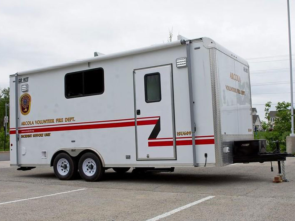 A white trailer labeled “ARCOLA VOLUNTEER FIRE DEPT. INCIDENT SUPPORT UNIT” is parked in a lot. It has emergency stripes, department logos, and two wheels on each side. Trees and pavement are visible in the background.