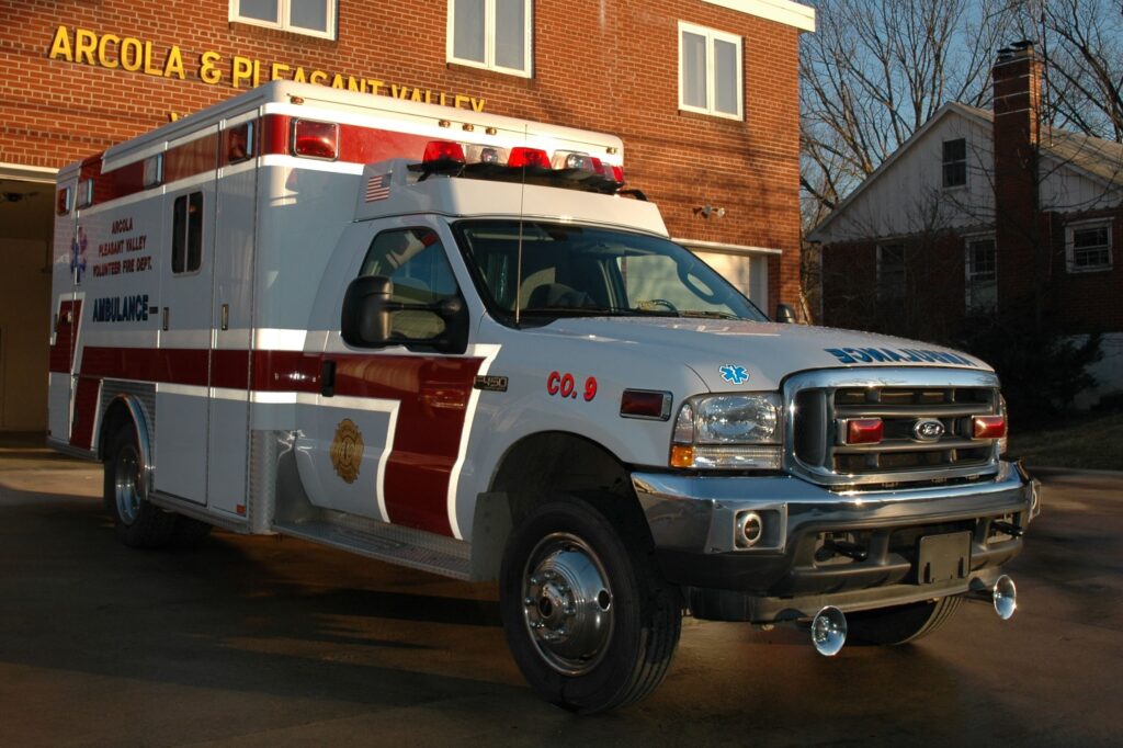 A white and red ambulance is parked outside a brick fire station, with the sun casting shadows. The vehicle has emergency lights on top and medical symbols on the side.