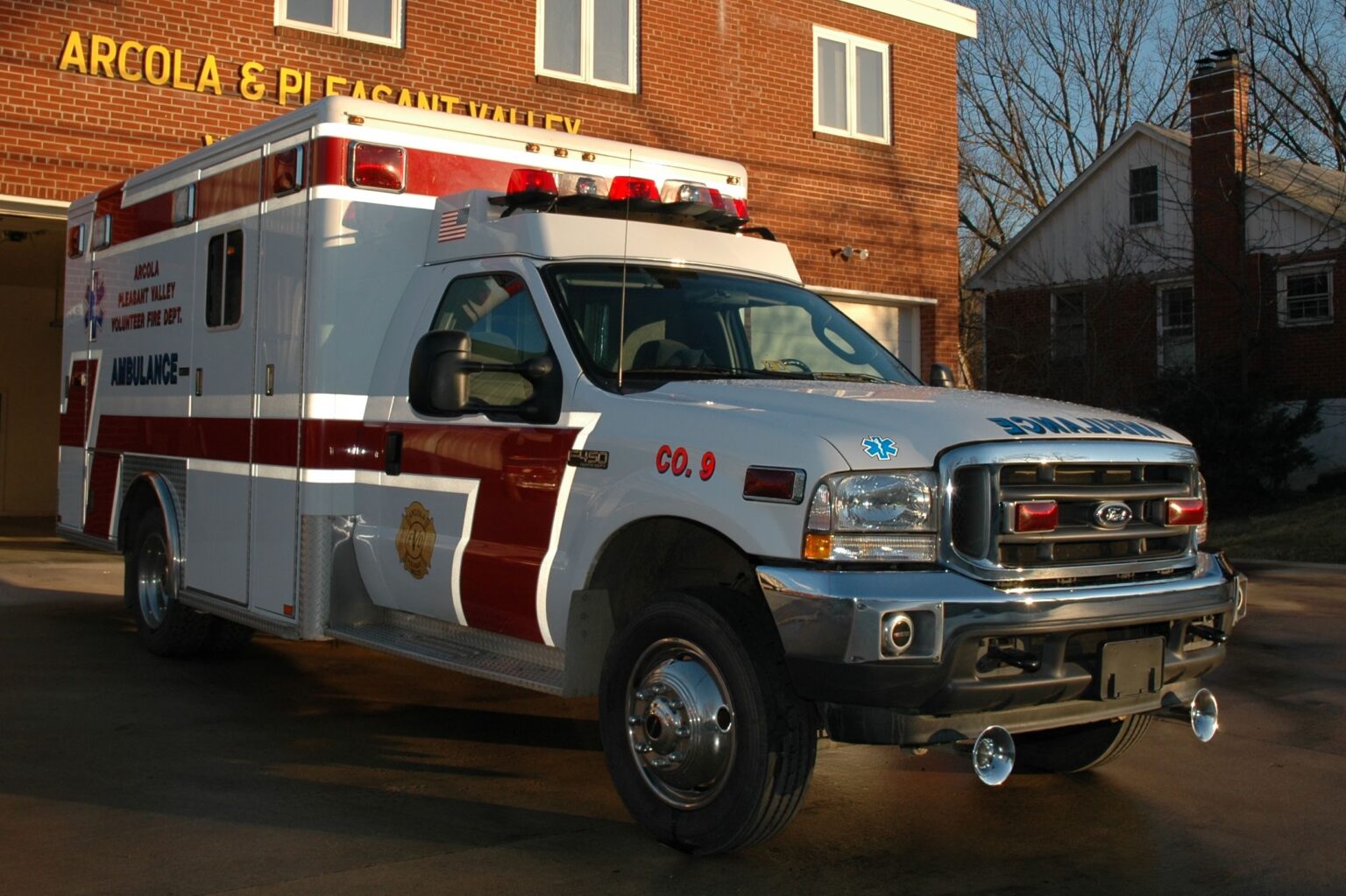 A white and red ambulance is parked outside a brick fire station, with the sun casting shadows. The vehicle has emergency lights on top and medical symbols on the side.