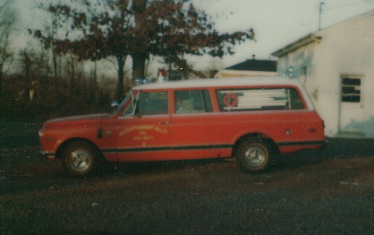 A vintage red emergency vehicle with a white roof is parked on grass near a white building and trees. The vehicle has emergency lights and a medical symbol on the side window.