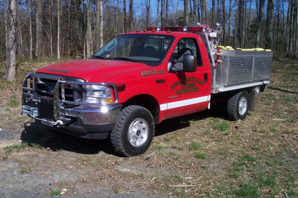 A red brush fire truck with a silver utility bed is parked on grass near trees. Equipment and hoses are stored on the truck bed. The truck says “Brush 655” and “Fire Department” on the side.