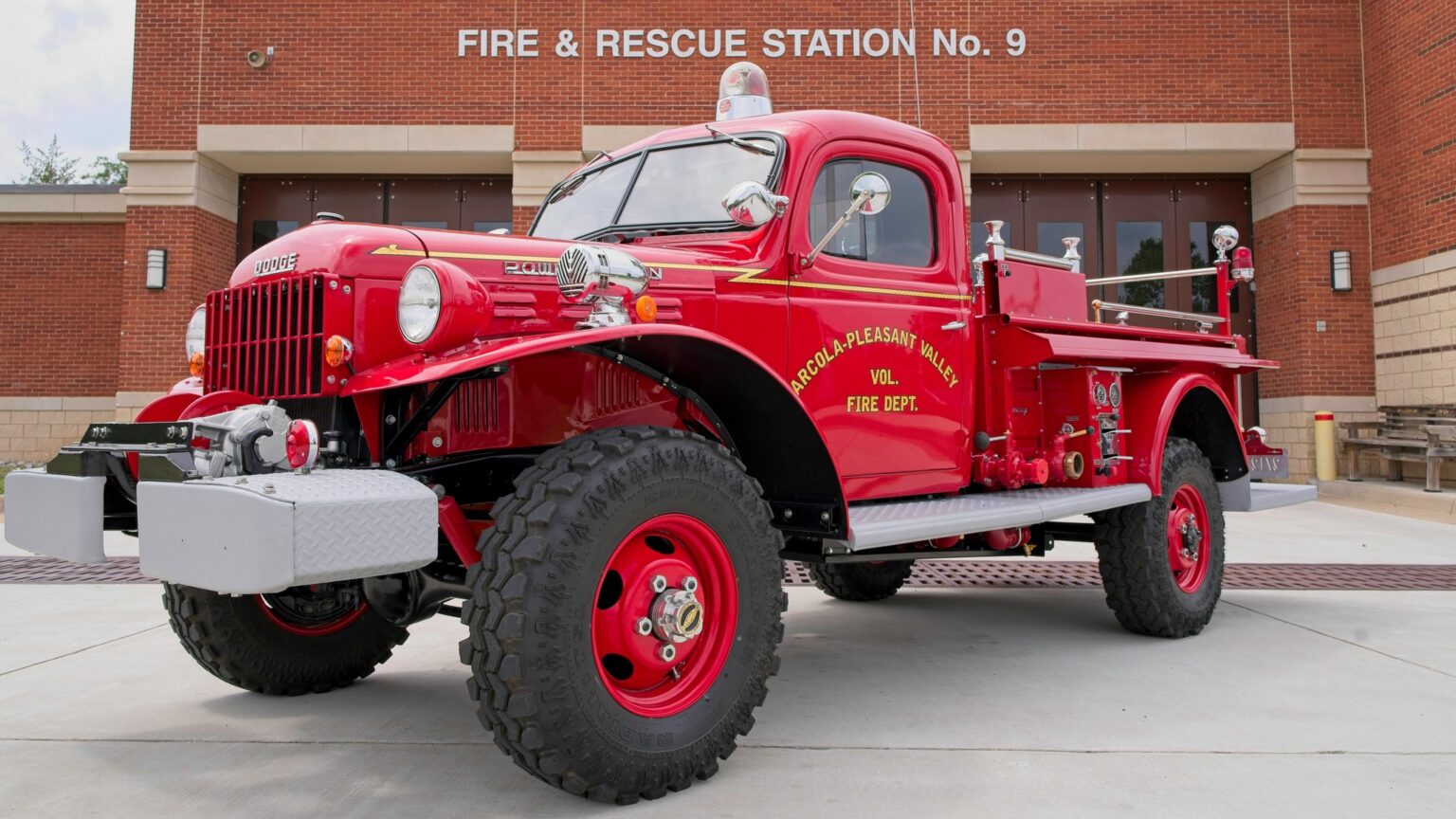 A vintage red fire truck is parked outside Fire & Rescue Station No. 9. The truck has large tires and shiny chrome details, and the fire station’s brick building is visible in the background.