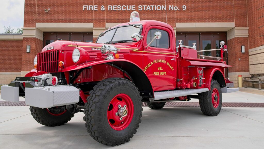 A bright red vintage fire truck is parked outside Fire & Rescue Station No. 9. The truck has large tires, chrome details, and is labeled “Arnold-Pleasant Valley Vol. Fire Dept.” in yellow script.