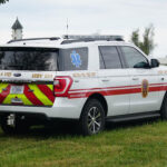 A white Arcola Volunteer Fire Department SUV with red stripes and emergency decals is parked on grass near a white fence and trees.