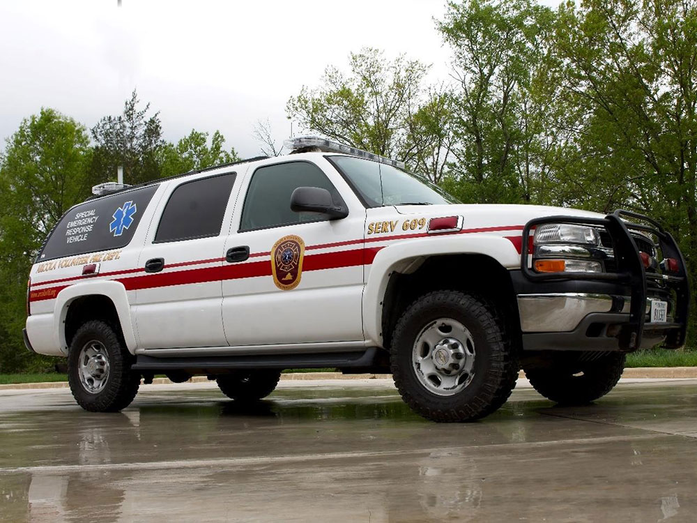 A white emergency response SUV with red stripes, an official badge, and a blue medical star parked on wet pavement, with trees in the background.
