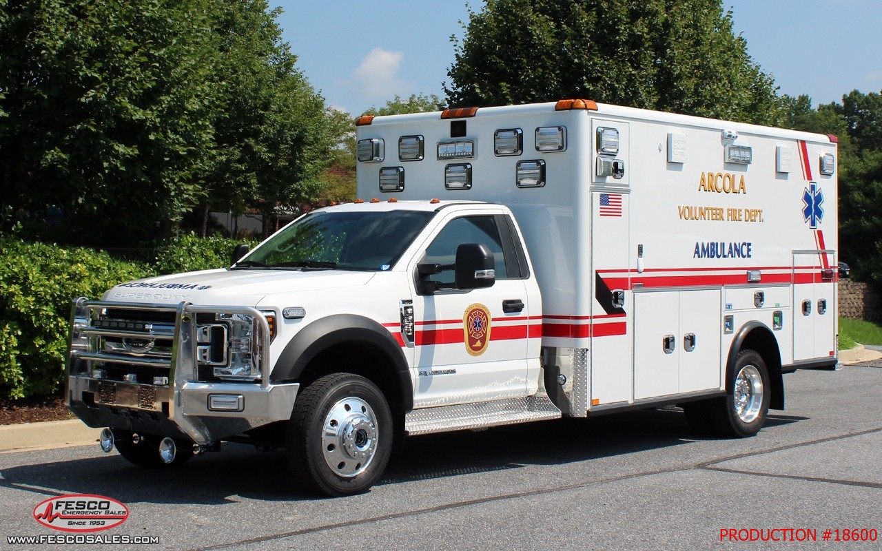 White ambulance truck with red stripes, labeled “ANOCLA VOLUNTEER FIRE DEPT.” parked outside on pavement, trees in the background, and emergency lights on top.