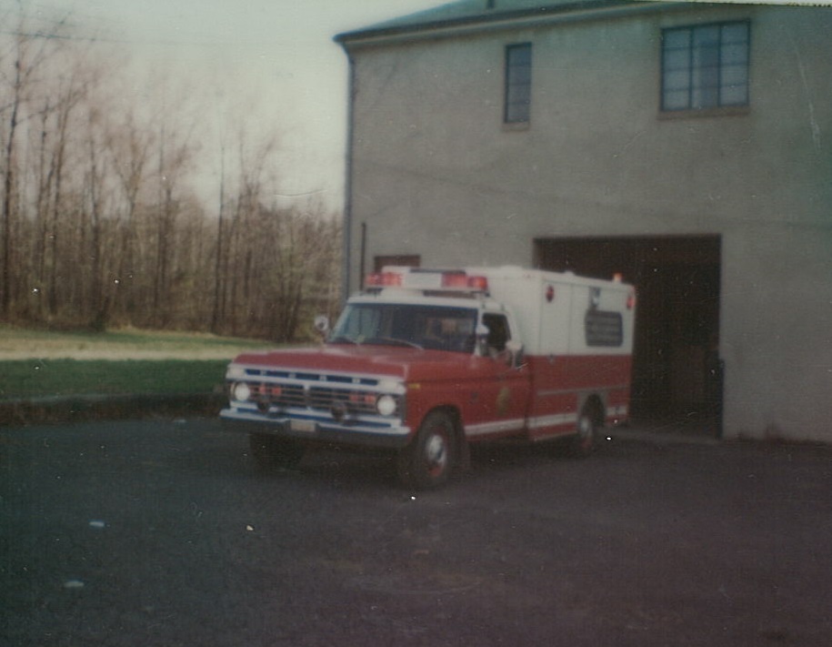 A vintage red and white ambulance is parked outside a beige building, with trees and grass visible in the background.