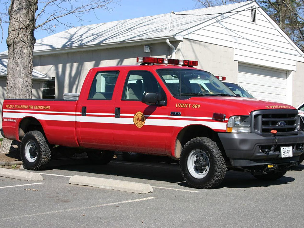 A red fire department utility truck is parked in a lot in front of a white building. The truck has emergency lights on top and fire department markings on the side.