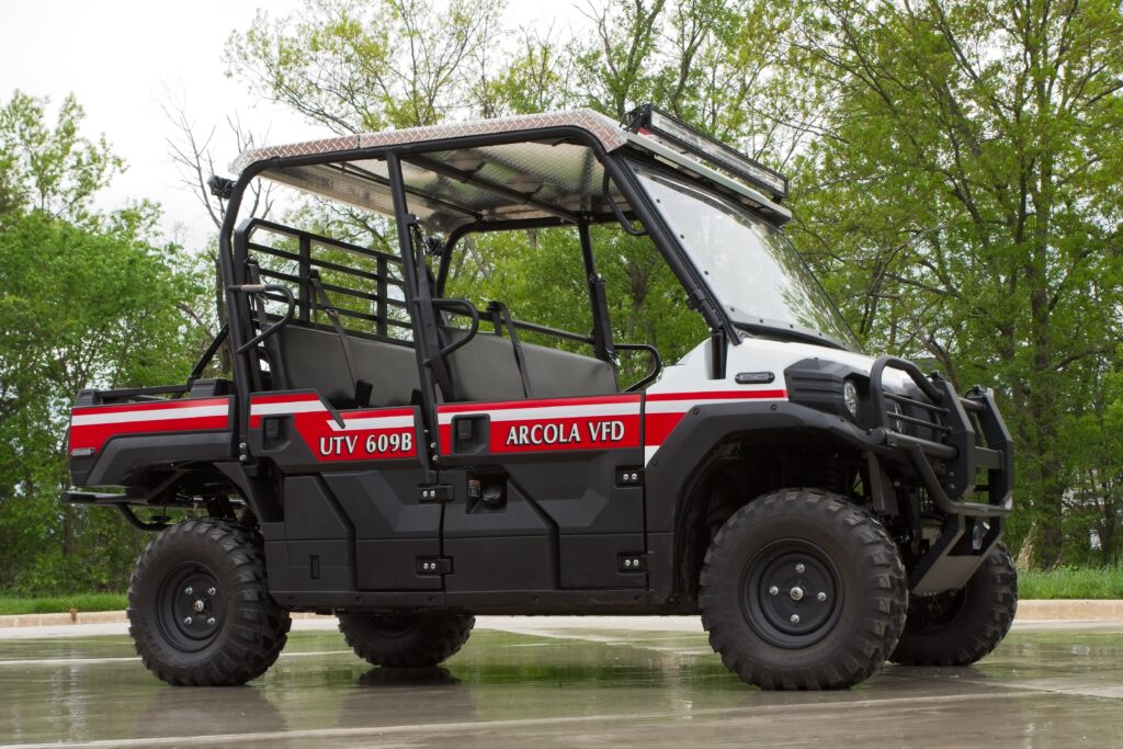 Black utility vehicle marked “UTV 609B” and “Arcola VFD” with red and white stripes, parked on wet pavement, surrounded by green trees in the background.