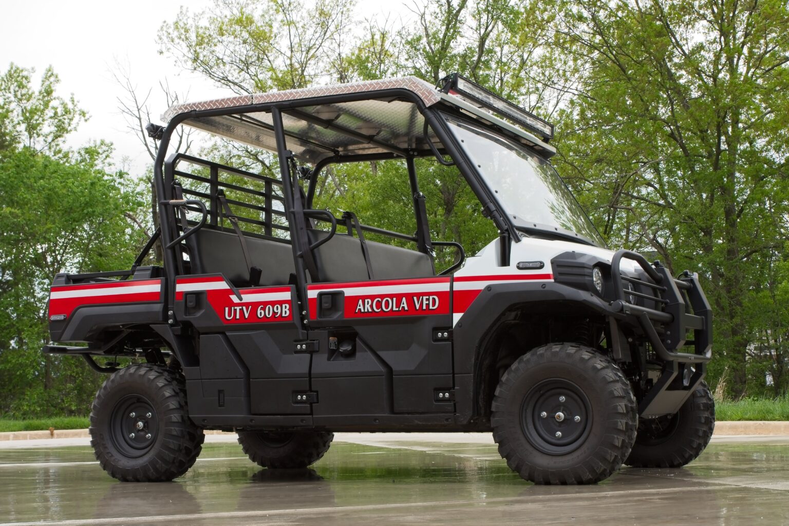Black utility vehicle marked “UTV 609B” and “Arcola VFD” with red and white stripes, parked on wet pavement, surrounded by green trees in the background.