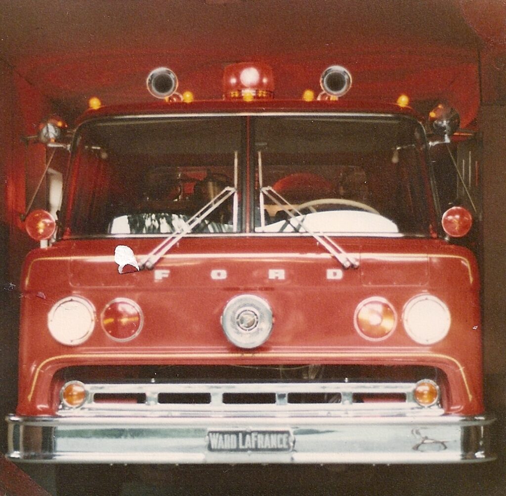 Front view of a vintage red Ford fire truck with “WARD LAFRANCE” on the license plate, two large headlights, and emergency lights on top, parked inside a garage.