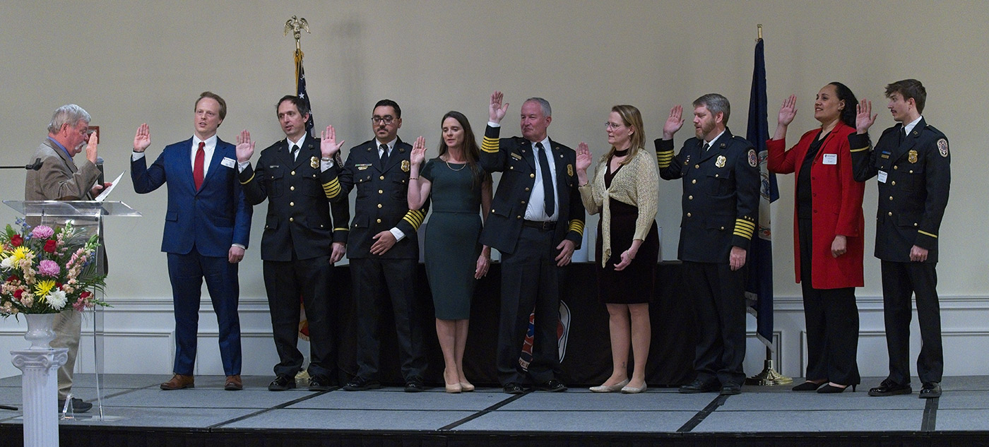 A group of nine people stands on a stage with their right hands raised, being sworn in by a man at a lectern; flags and flowers are visible in the background.