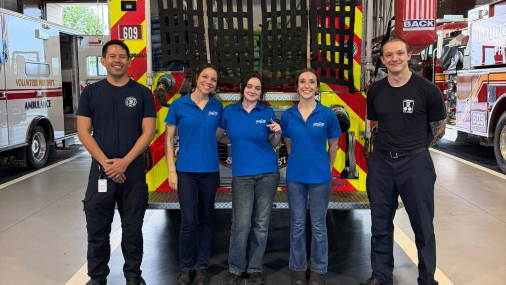 Five people stand smiling in front of a firetruck inside a fire station. Three women wear blue polo shirts, while two men wear dark shirts and pants. Emergency vehicles are visible on each side.