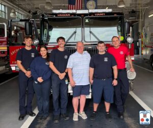 Six people stand smiling in front of a Loudoun County fire truck inside a fire station. Five wear firefighter uniforms; one wears a striped polo and shorts. The Loudoun County Fire & Rescue logo is in the lower right corner.