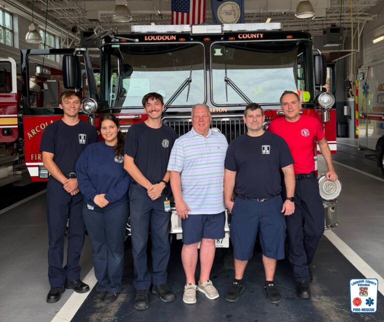 Six people stand smiling in front of a Loudoun County fire truck inside a fire station. Five wear firefighter uniforms; one wears a striped polo and shorts. The Loudoun County Fire & Rescue logo is in the lower right corner.