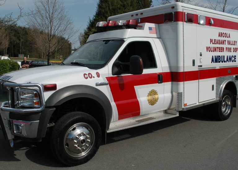 A white and red ambulance from Arcola Pleasant Valley Volunteer Fire Dept. is parked on a street, with emergency lights on its roof and medical symbols on the side.