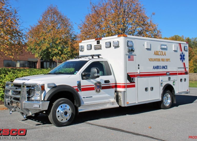 A white Alscola Volunteer Fire Dept. ambulance is parked on a street near trees with autumn leaves and a brick building in the background. The vehicle has red stripes and emergency markings.