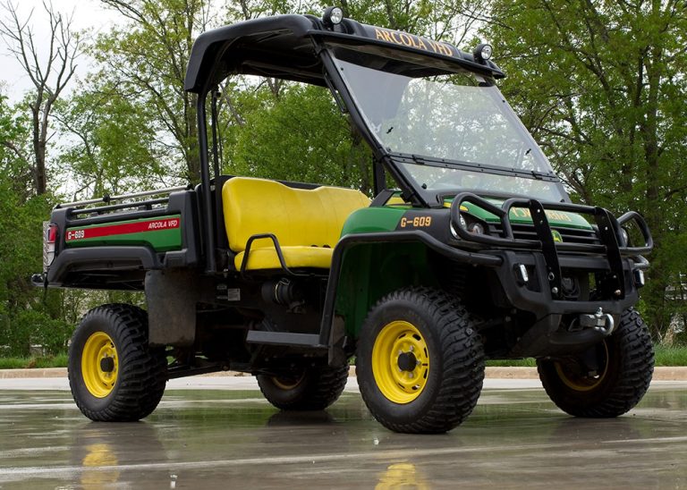 A green and yellow utility vehicle with large tires and an open cab, parked on wet pavement with trees in the background.