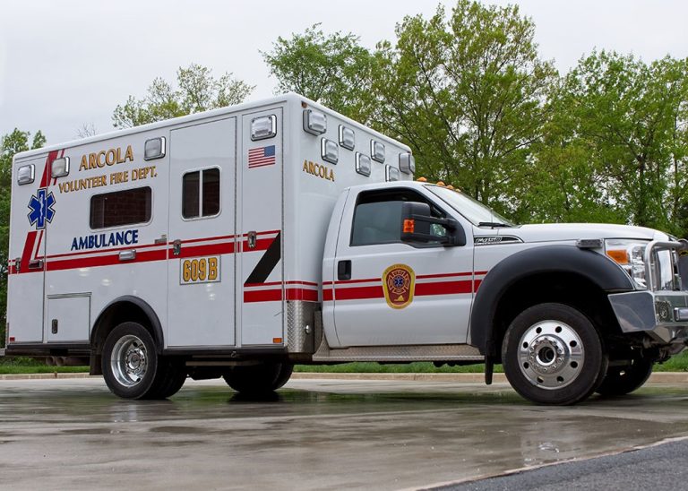 A white Arcola Volunteer Fire Dept. ambulance is parked on wet pavement, with trees and cloudy sky in the background.